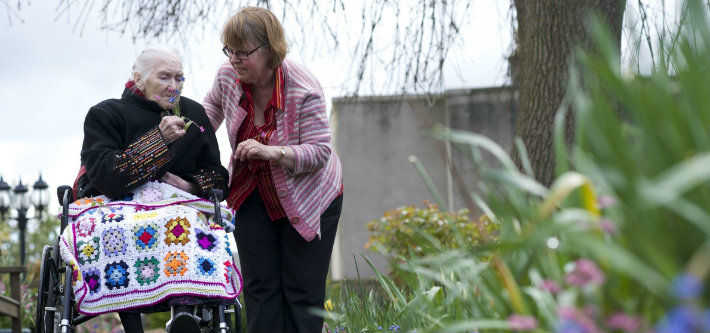 an elderly lady sitting in a wheelchair with a crochet blanket over her lap with her carer outside 