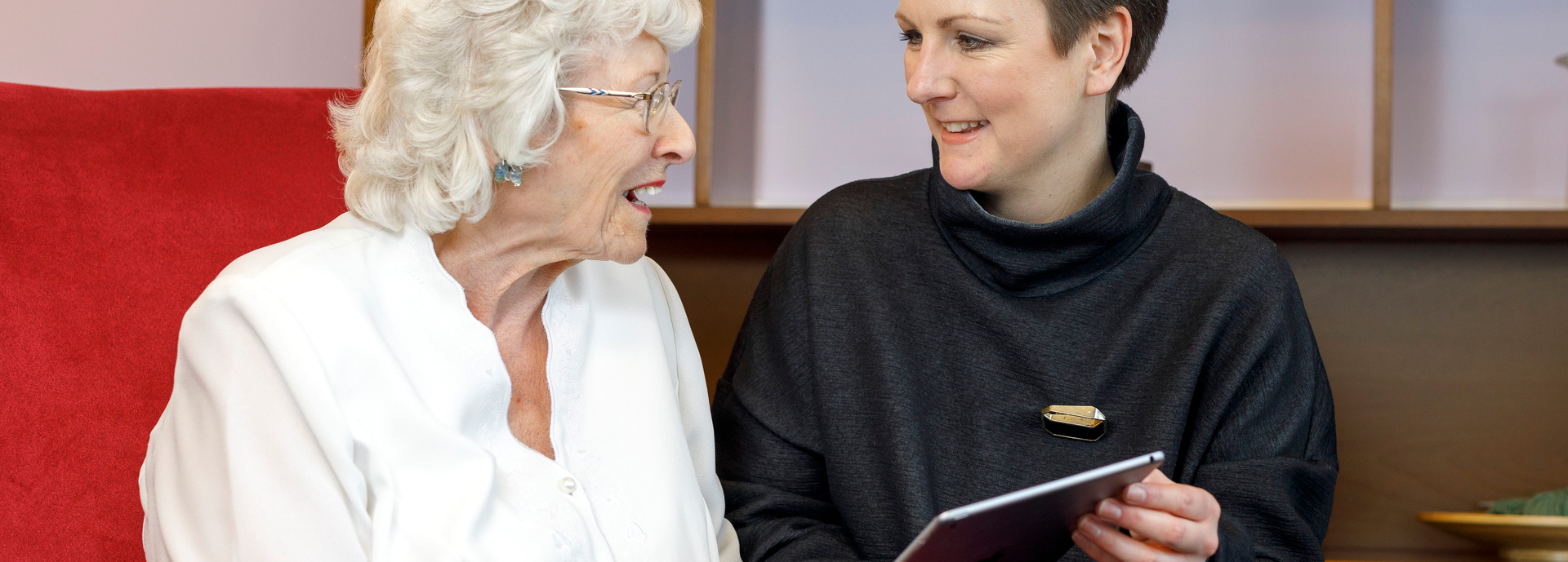 Two women sat next to each other. The younger one is showing the other elderly lady something in a book.