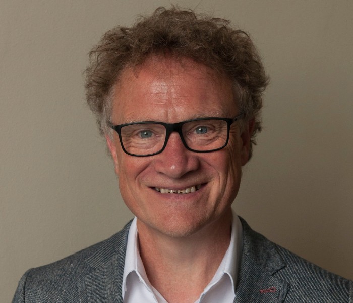 Prof Adrian Blackridge looks straight to camera, smiling. The photo shows his head and shoulders only. He wears black, square glasses and the top of a white shirt and grey blazer can be seen.