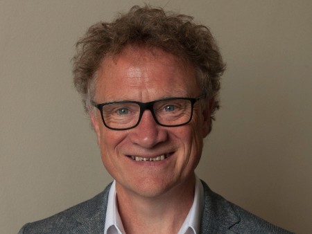 Prof Adrian Blackridge looks straight to camera, smiling. The photo shows his head and shoulders only. He wears black, square glasses and the top of a white shirt and grey blazer can be seen.