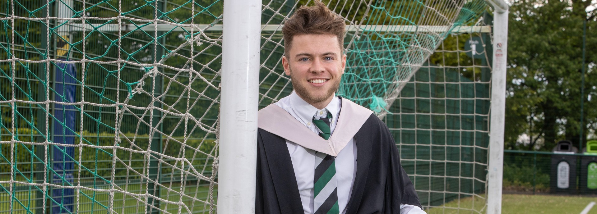 Footballer Craig Brown standing against a goalpost wearing his graduation robes