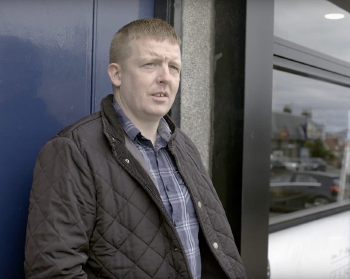 Malky - a white male with light coloured hair wearing a blue shirt and quote, is standing in a doorway