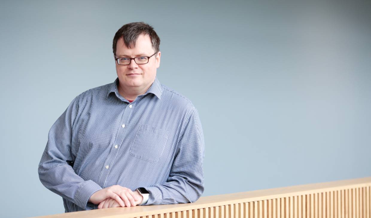 Photograph of academic Grant Gibson. A man looks straight to camera. He has dark rimmed square glasses on, dark short hair and is wearing a blue shirt.