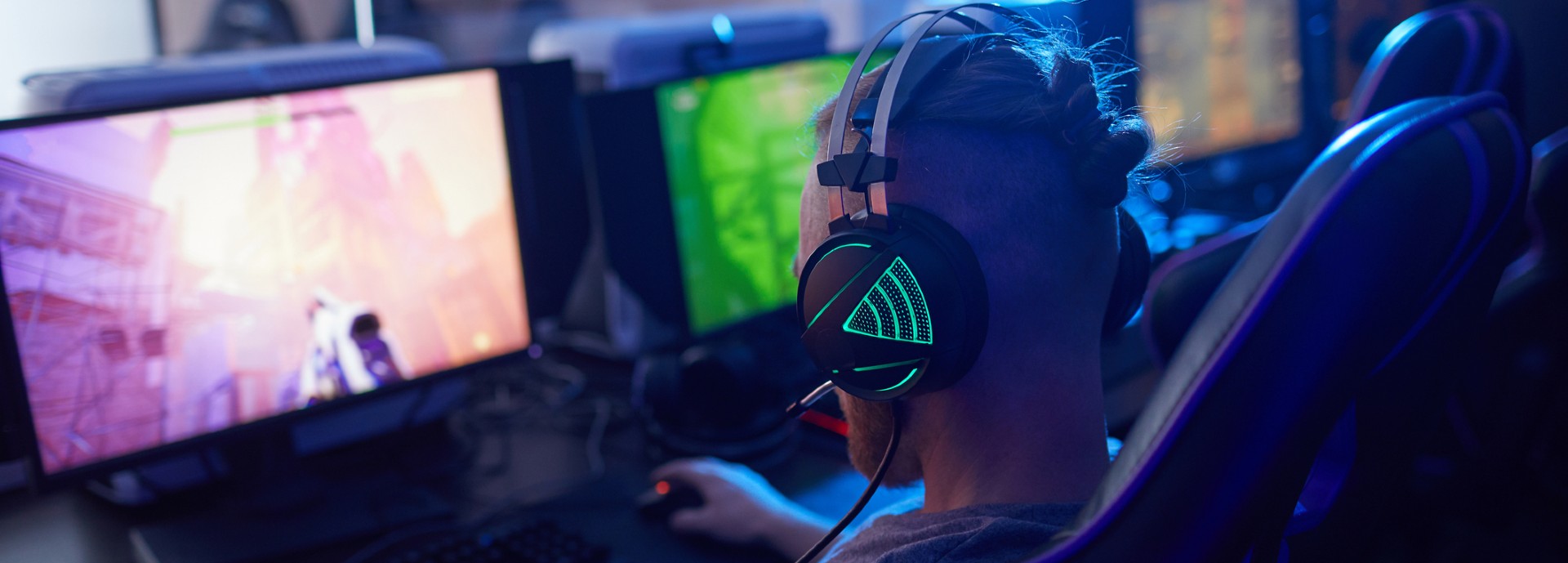 Rear view of young gamer wearing gaming headphones with backlight and playing in computer video game on computer