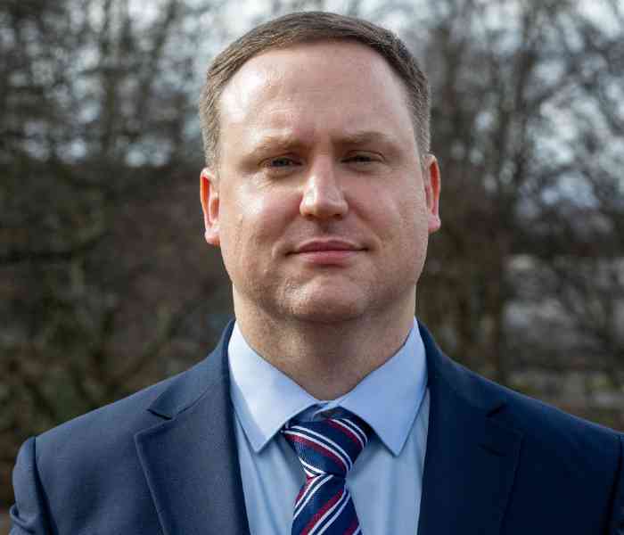 A photograph of Dr Daniel Jones, a white male dressed in a shirt, tie and suit jacket