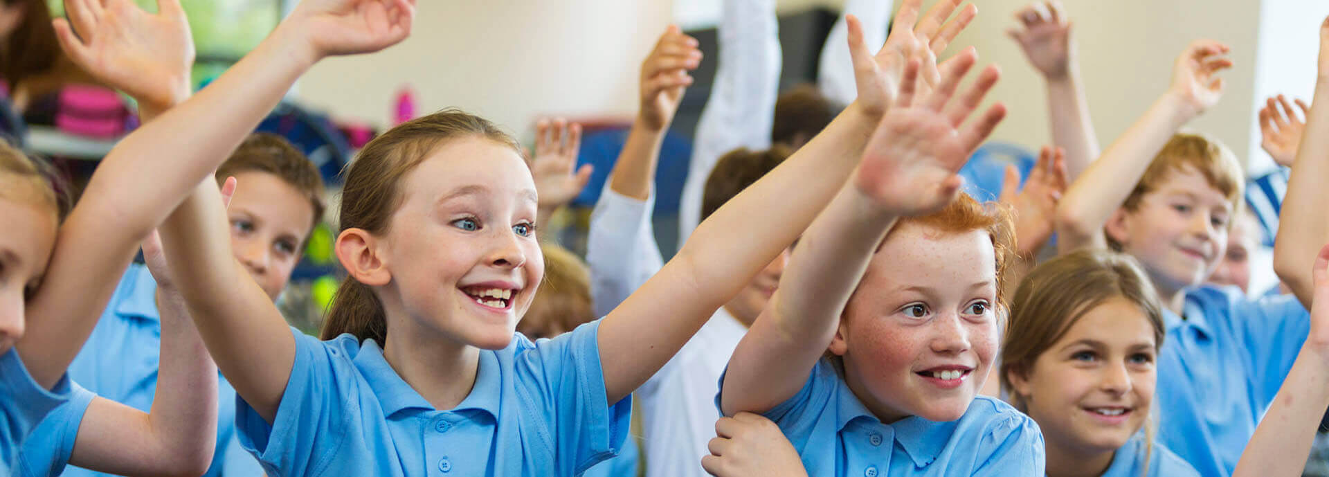 Excited school children in school uniform with hands up ready to answer a question from the teacher