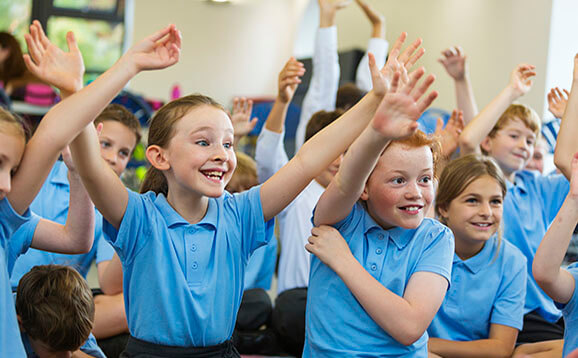 Excited school children in school uniform with hands up ready to answer a question from the teacher