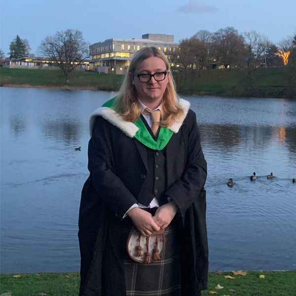 Lewis stood by the loch wearing a graduation gown